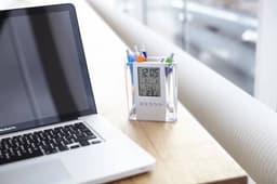 A transparent ABS pen holder with clock containing various pens, placed next to a silver laptop on a wooden surface.