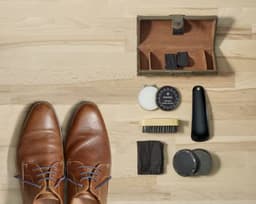 A brown shoe polish set displayed on a wooden surface, including brushes, polish, cloth, and a storage case.