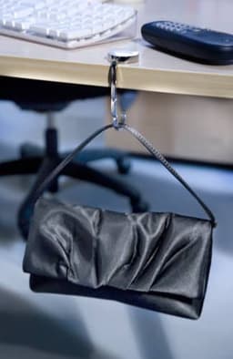 A black metal bag hook holding a textured black clutch bag under a desk, with a computer keyboard and a remote nearby.