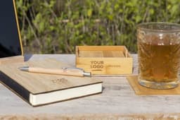A bamboo coaster set with a matching pen and notebook, accompanied by a glass of drink on a wooden surface.