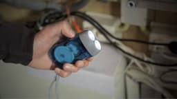 A blue ABS dynamo torch with three bright LEDs, held in a hand, against a background of cables and equipment.