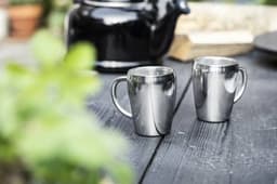Two stainless steel double-walled mugs sit on a wooden table, with a black kettle and greenery in the background.