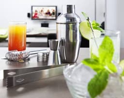 A stainless steel cocktail set featuring a shaker, jigger, strainer, tongs, and glasses with colourful beverages.