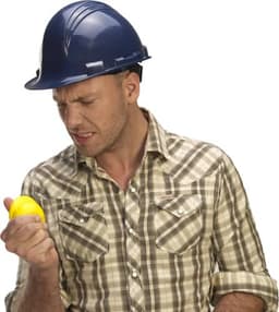A man in a blue hard hat holds a yellow stress ball, displaying a frustrated expression.