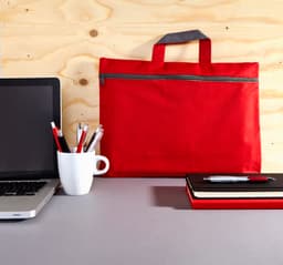 A red polyester conference bag with a grey handle, placed on a grey desk beside a laptop, pens, and notebooks.