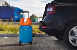 A blue wheeled trolley bag with an orange handle, positioned beside a car on a paved surface.