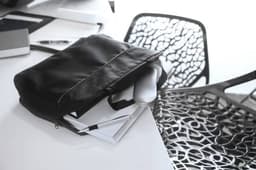 A black polyester document bag lying on a desk, surrounded by stationery and a stylish chair.