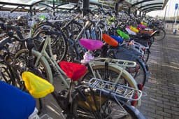 Colourful polyester bicycle seat covers in bright pink, red, yellow, green, and blue are fitted on bikes in a rack.