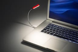 A red and silver laptop light is attached to a silver laptop, illuminating the keyboard. The background is dark.