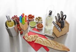 A wine bottle in a cooler, colourful utensils, fresh ingredients, a cutting board, and various knives on a grey surface.