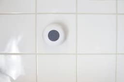 A round white ABS speaker with a grey control panel on a tiled white surface.