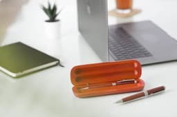 A rosewood ballpen in a wooden case, placed on a desk near a laptop, notepad, and a small plant.