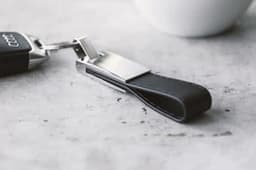 A black leather and silver key holder sits on a concrete surface beside a white cup. It features a logo.