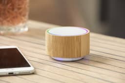 A bamboo speaker with a circular white top, sitting next to a smartphone on a wooden surface.