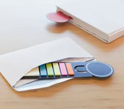 Cardboard sticky note holder with colourful sticky notes and a circular section, placed inside an envelope on a wooden surface.