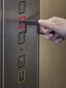 A black aluminium door opener is being used to press the red elevator button.