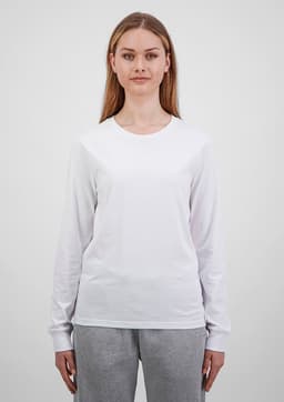 A woman wears a white long sleeve tee, paired with grey trousers. The tee features a simple design and has a logo.