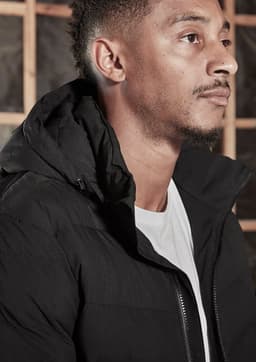 A black hooded puffer jacket, featuring a zipper and a discreet logo, worn by a model.