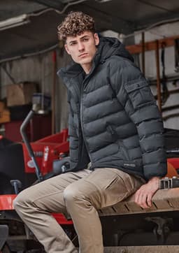A unisex hooded puffer jacket in black, featuring a fitted design and a logo, worn by a model sitting on machinery.