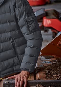 A grey unisex puffer jacket with a zip and pocket detail on the sleeve, displayed on a person near machinery.