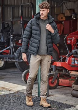 A black hooded puffer jacket is worn over a grey shirt, paired with beige pants and tan boots in an outdoor setting.