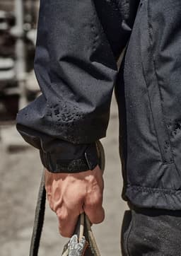 A close-up of a person's arm wearing a black packable rain jacket, holding a rope. The jacket features an adjustable cuff.