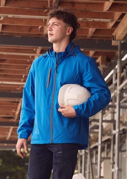 A man wears a blue unisex packable rain jacket, holding a white helmet, set against a construction backdrop.