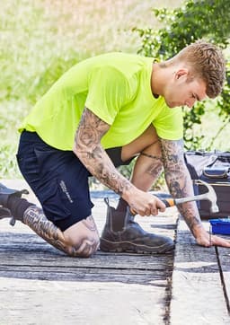 A person kneeling outdoors in bright yellow clothing and dark blue sports shorts, using a saw on a wooden surface.