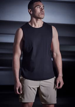 A man wearing a black sleeveless top and beige sports shorts stands in a dimly lit setting. The shorts feature a logo.