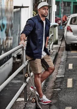 A man wearing a navy shirt and beige stretch shorts stands by a railing, with a skateboard beside him.