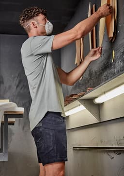 A man wearing a light grey shirt and dark shorts is organising materials on a wall in a workshop.