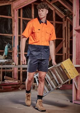 A man wearing an orange and navy work shirt and navy shorts carries a ladder in a construction setting.