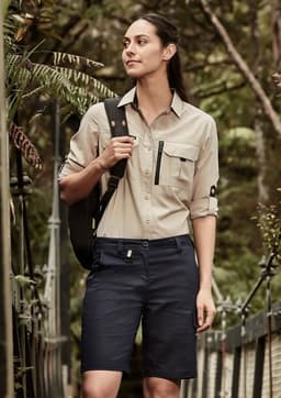 A woman in a light beige shirt and navy shorts stands outdoors, with a backpack over her shoulder. The shorts feature a logo.