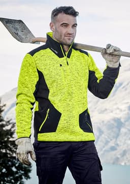 A man holds a shovel while wearing a bright yellow and black reinforced knit 1/2 zip pullover, set against a mountainous backdrop.
