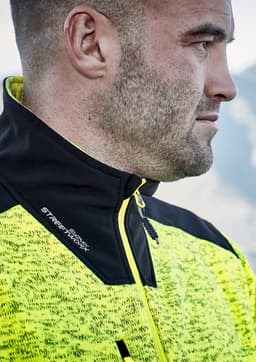 A close-up of a man wearing a bright yellow and black reinforced knit 1/2 zip pullover, featuring a branding logo.