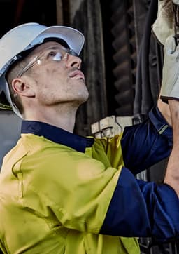 A man in a yellow and navy long sleeve shirt with a helmet and safety glasses, examining equipment.