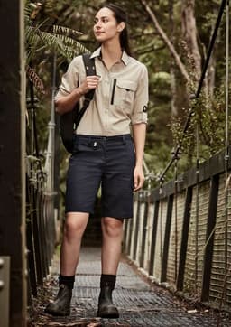 A woman in a beige long sleeve button-up shirt and navy shorts stands on a forest walkway, carrying a bag.
