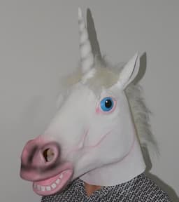 A unicorn mask in white with a pink snout, blue eyes, and a horn, worn by a person against a plain background.