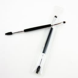 A double-headed eyebrow brush with a black grip, featuring a spoolie on one end and an angled brush on the other, in clear packaging.