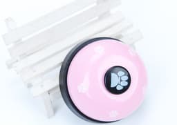 A pink pet training bell with a black centre, featuring paw print designs, placed on a white wooden stand.