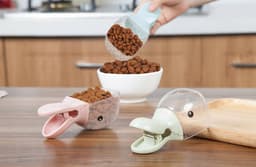A pet food scoop in pink and green, with a clip, above a bowl of kibble on a wooden surface.