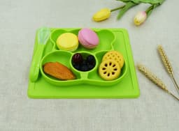 A green non-slip children's plate with compartments holding various treats, including macarons, fruit, and a cookie.