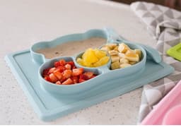 A blue, non-slip children's plate with three sections, holding strawberries, bananas, and pineapple on a countertop.
