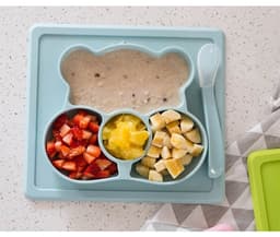 A blue non-slip children's plate shaped like a bear, featuring sections for fruit and porridge, with a matching spoon.