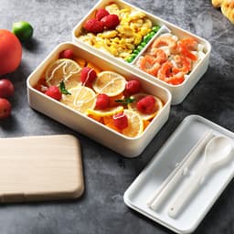 A double-layer lunch box set in white containing fruits, vegetables, scrambled eggs, and seafood, with a wooden lid and utensils.