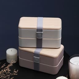 A double-layer lunch box set in cream and taupe, secured with a grey strap, placed near candles.