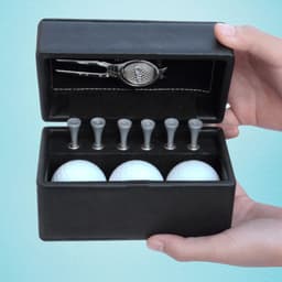 A black golf set box containing three white golf balls, six wooden tees, and a silver divot tool.