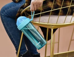 A drink bottle in turquoise with a compartmentalised double drinking straw, held by a person sitting on a gold chair.