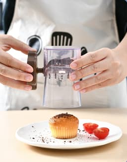 A manual mashing and grinder in clear plastic with a brown handle, used above a plate with a cupcake and tomatoes.