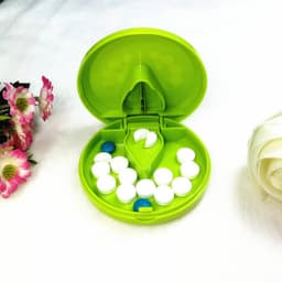 A green plastic pill cutter and pill box, containing white and blue pills, with floral decorations nearby.